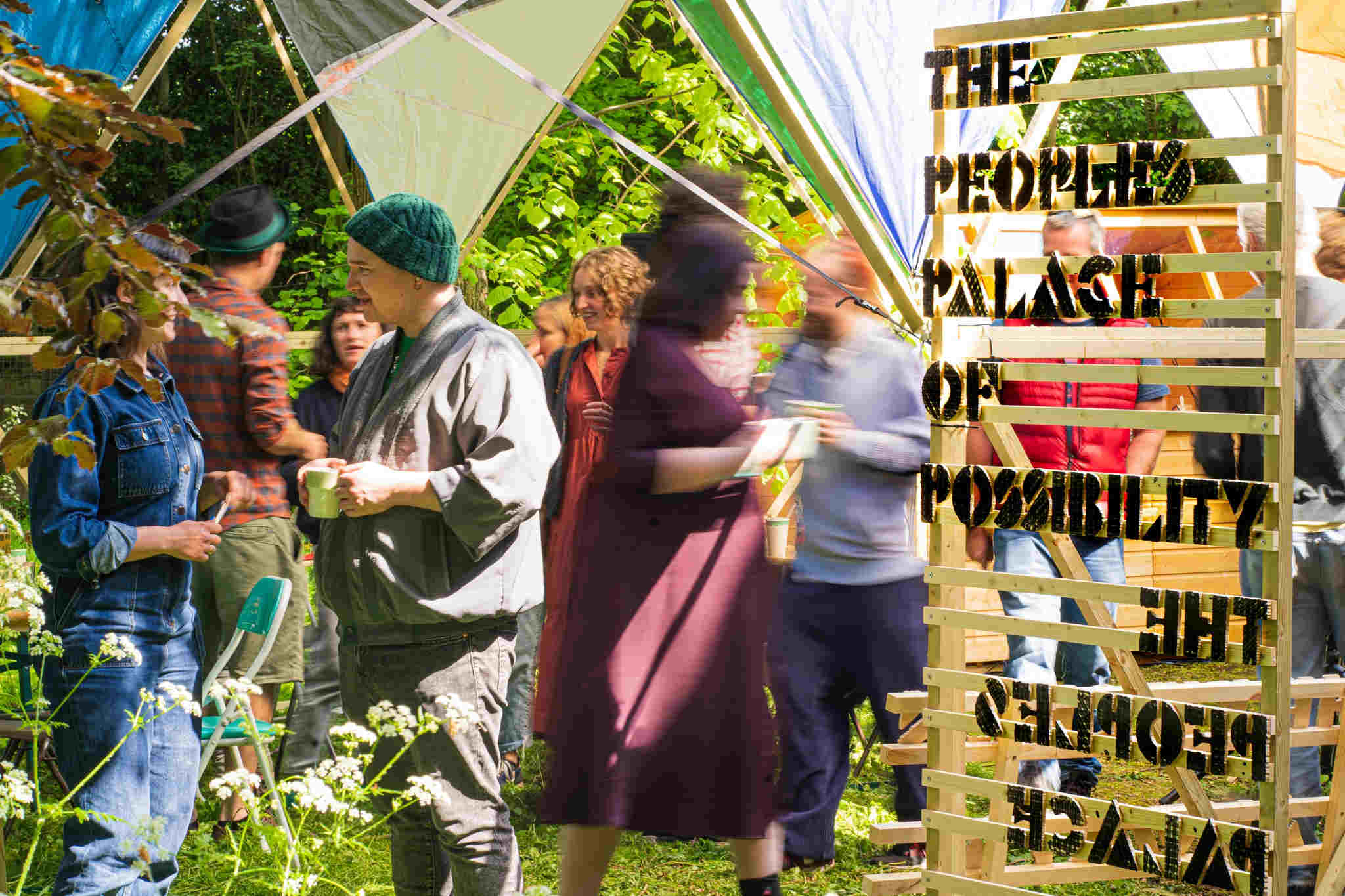 A door frame leading into a space with people walking inside The People's Palace - a geodesic dome. The people are blurred. A wooden door frame with slats leads into the space and has text on it which reads The People's Palace of Possibility. There is grass in the foreground and trees in the background.